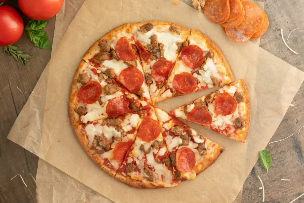 Top down photo of a baked Gluten Free Sausage & Pepperoni pizza. The pizza is on brown parchment paper with ingredients scattered all around.