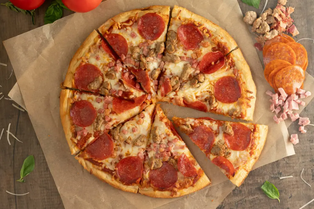 Top down photo of a baked Meat Lover's pizza. The pizza is on brown parchment paper with ingredients scattered all around.