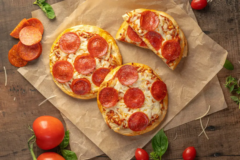 Top down photo of three baked mini Pepperoni pizzas. The pizzas are on brown parchment paper with ingredients scattered all around.