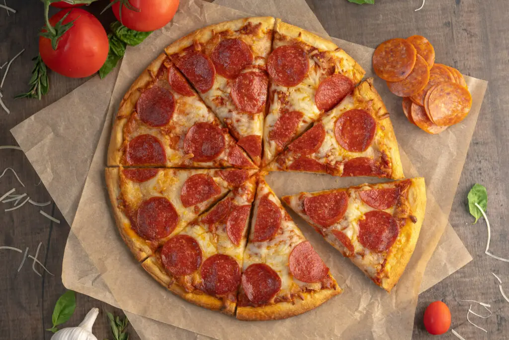 Top down photo of a baked Pepperoni pizza. The pizza is on brown parchment paper with ingredients scattered all around.