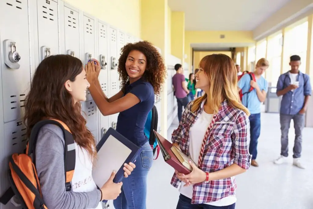 Three high school girls at their lockers in a school hallway. Other kids are in the background.