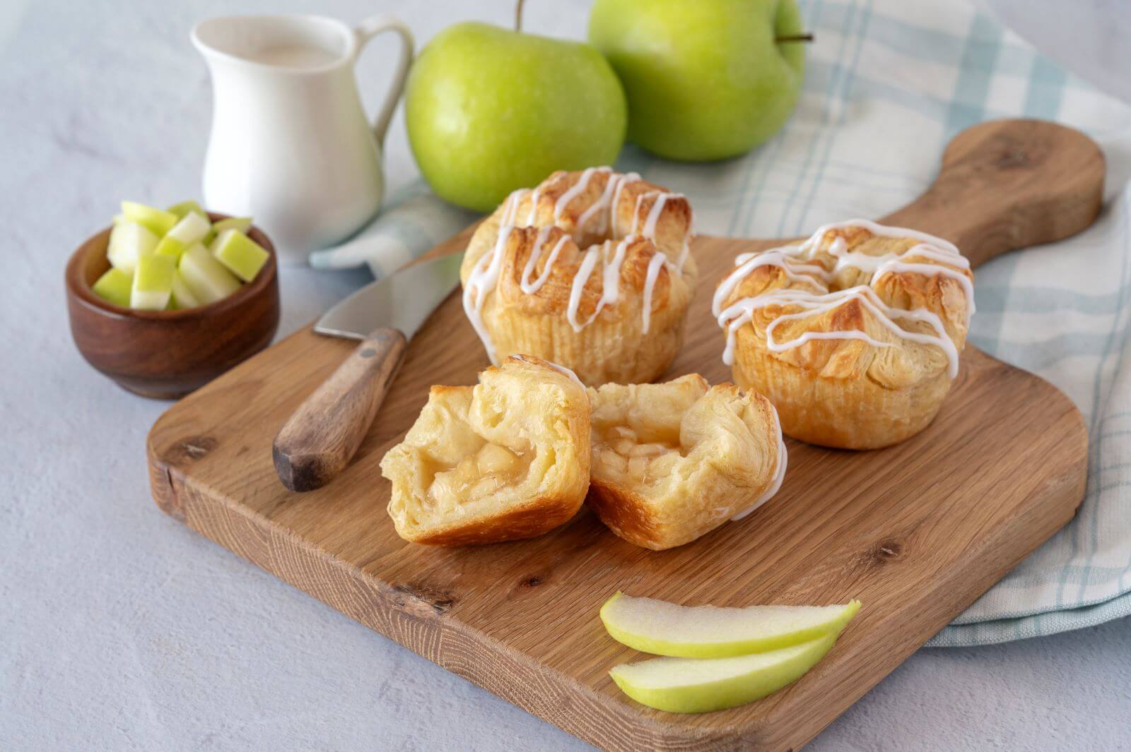 Three Apple Croissant Crowns on a wood cutting board. One of the crowns is cut in half. Around the crowns are slices of green apples, a bowl of green apple pieces, whole green apples, and a creamer.