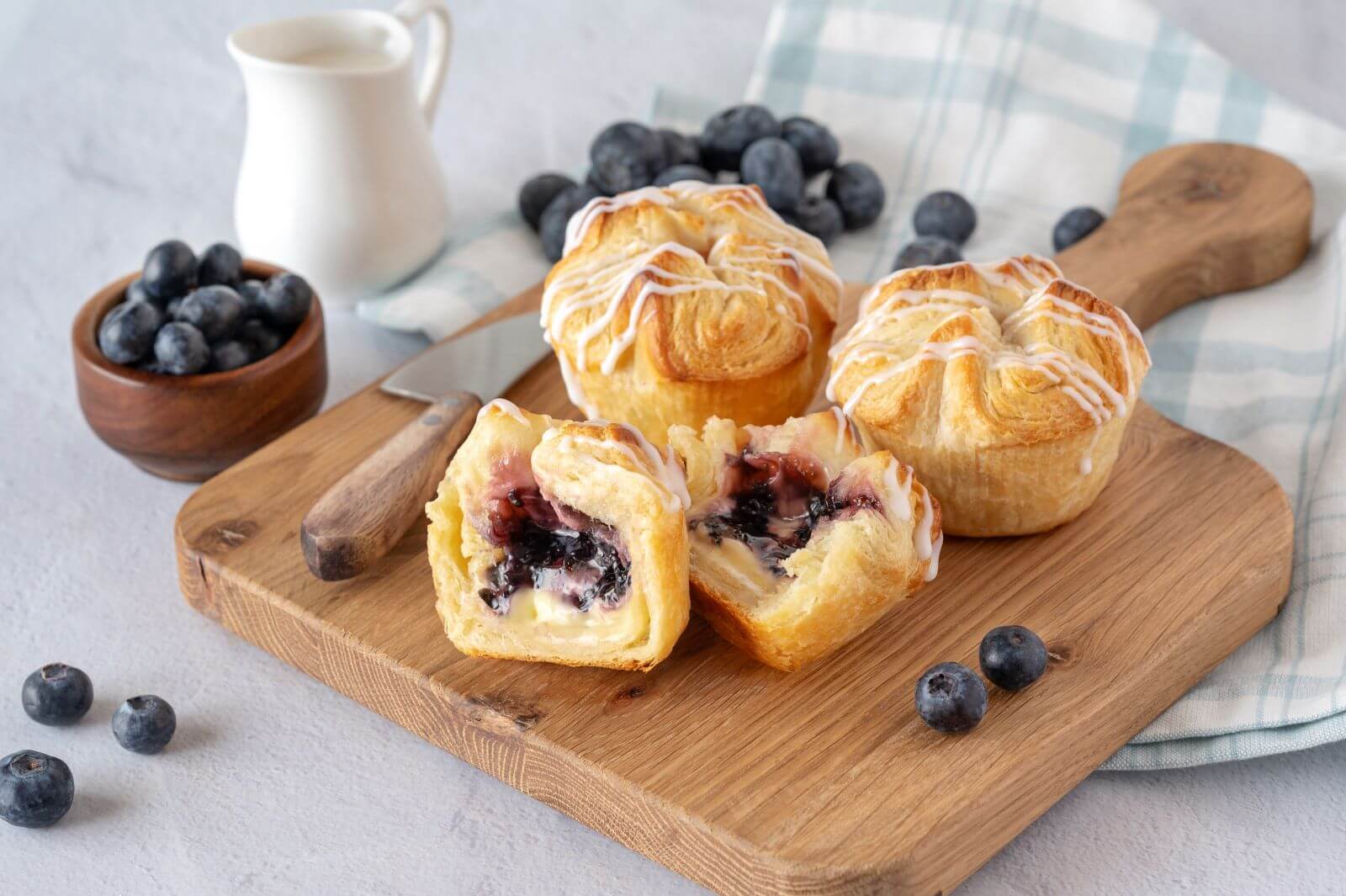 Three Blueberry & Cream Croissant Crowns on a wood cutting board. One of the crowns is cut in half. Around the crowns are blueberries, a bowl of blueberries, and a creamer.