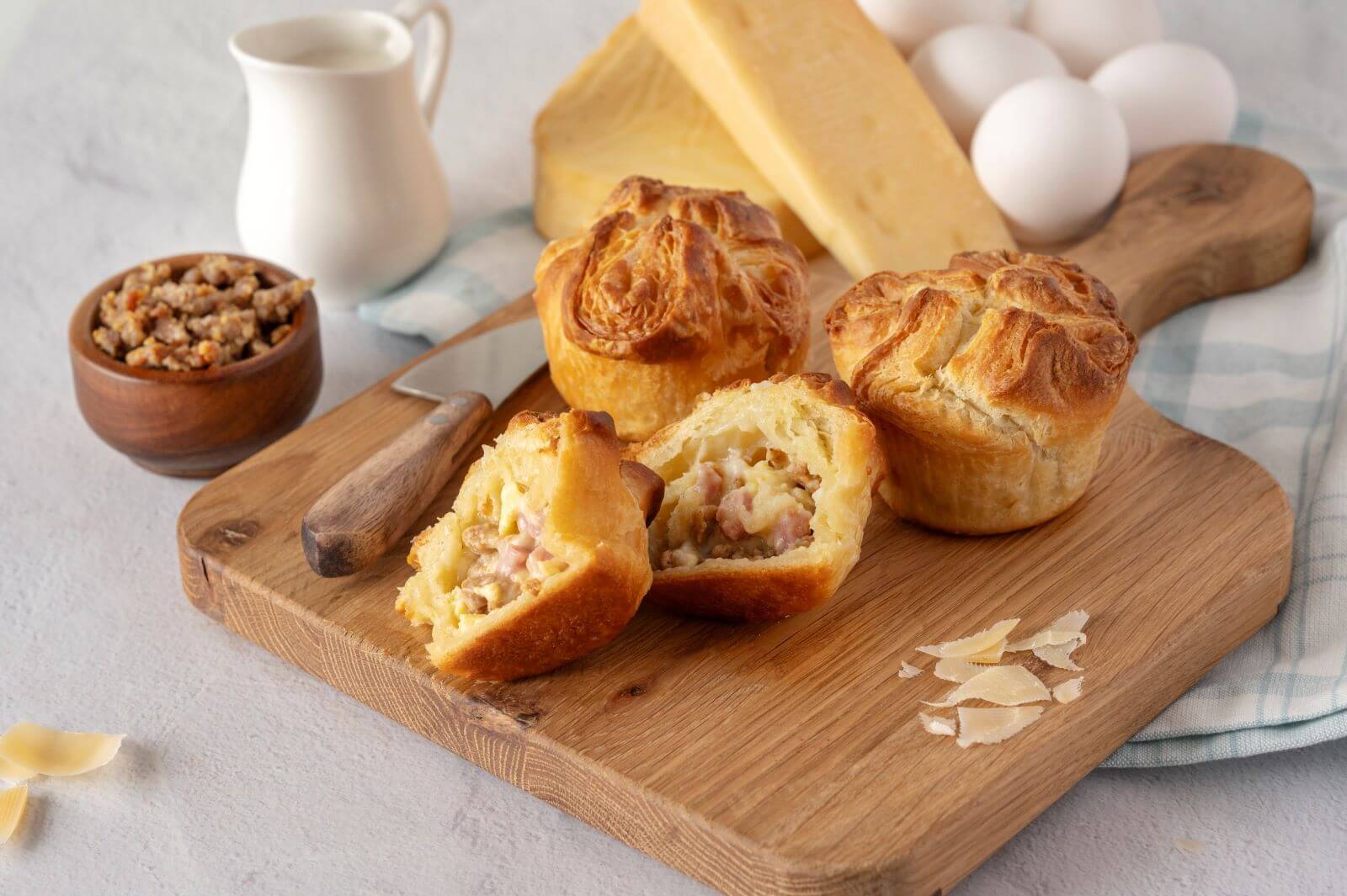 Three Breakfast Croissant Crowns on a wood cutting board. One of the crowns is cut in half. Around the crowns are slices of cheese, a bowl of cooked sausage, and a creamer.