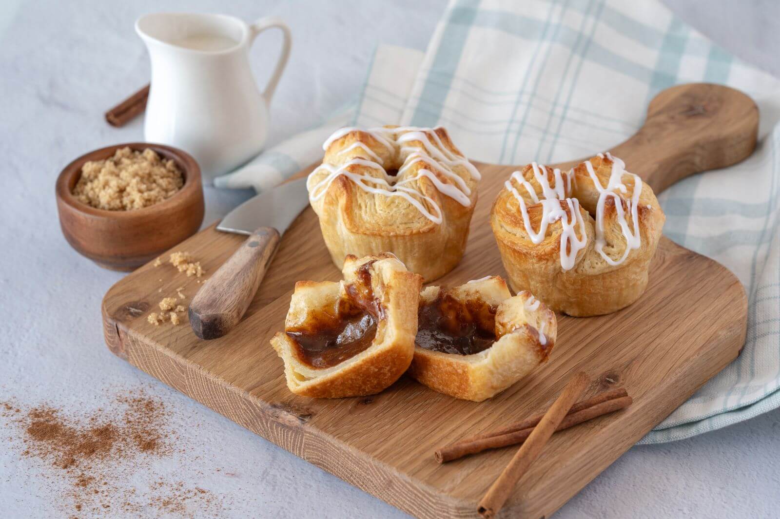 Three Cinnamon Croissant Crowns on a wood cutting board. One of the crowns is cut in half. Around the crowns are cinnamon sticks, a bowl of brown sugar, and a creamer.