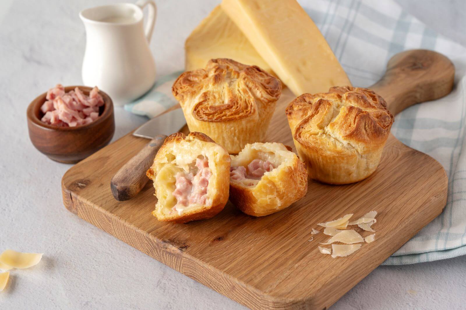 Three Ham & Cheese Croissant Crowns on a wood cutting board. One of the crowns is cut in half. Around the crowns are slices of cheese, a bowl of cubed ham, and a creamer.
